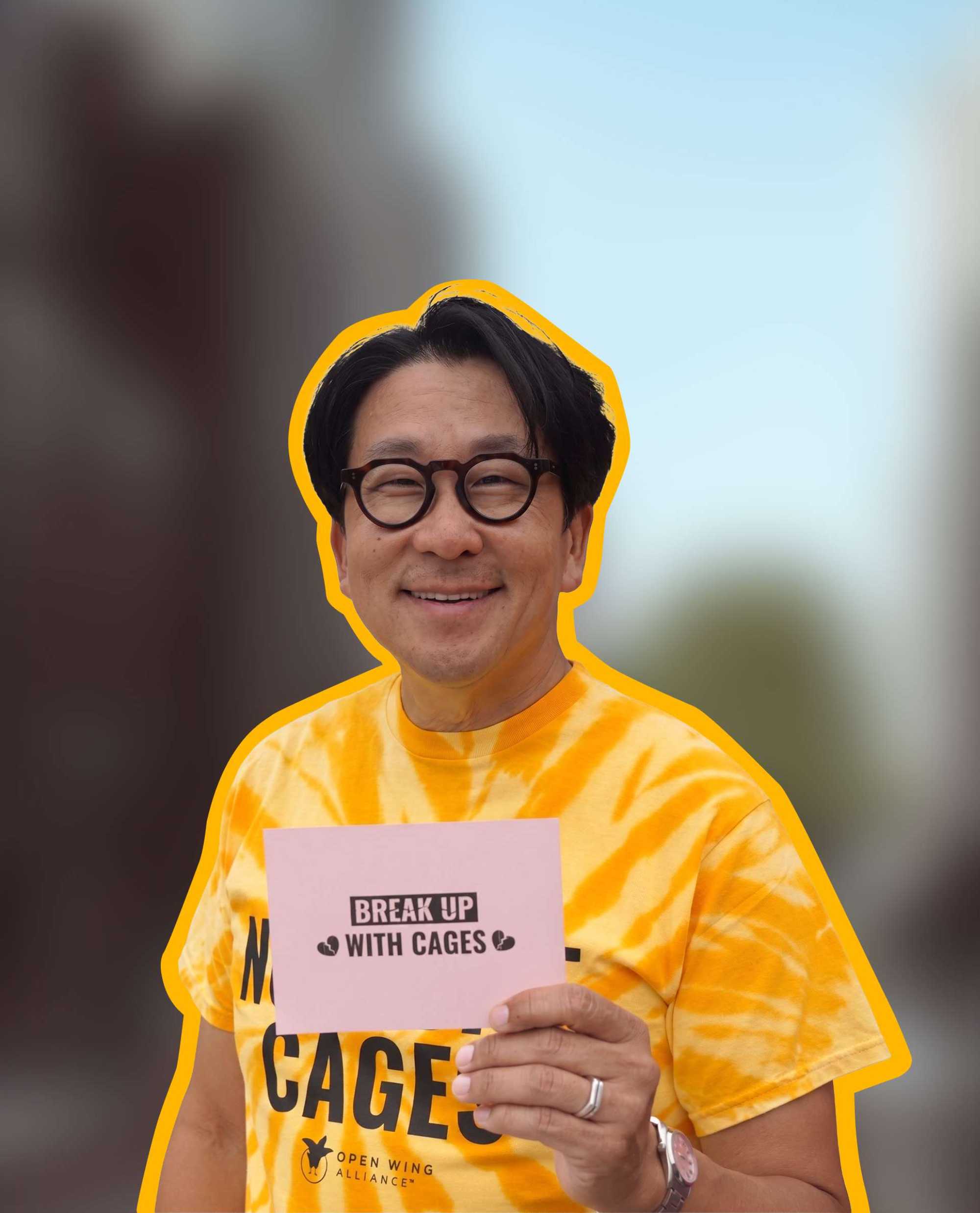 Nick Cho, a man with black hair and glasses, wears a yellow tie-die t-shirt and holds a sign that reads "BREAK UP WITH CAGES."